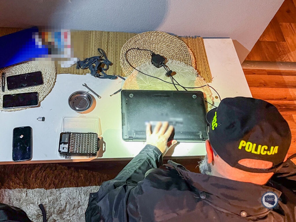Policjant zabezpiecza komputer. Wokół komputera znajdują się okablowanie oraz nośniki komputerowe.