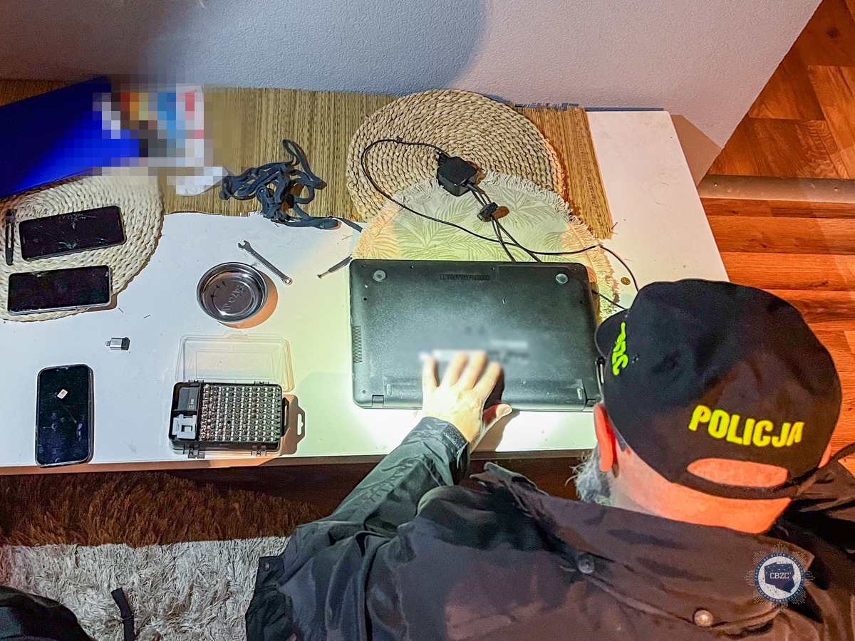 Policjant zabezpiecza komputer. Wokół komputera znajdują się okablowanie oraz nośniki komputerowe.